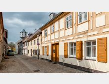 Schmale, gepflasterte Straße mit Fachwerkhäusern, die helle Fassaden und gelbe Fensterläden aufweisen. Im Hintergrund ist ein Wasserturm und mehrere Bäume zu sehen, während ein Auto am Straßenrand parkt.