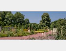 Ein gepflegter Garten mit geschwungenen Wegen, bunten Blumenbeeten und einzelnen Sträuchern, umgeben von hohen Bäumen im Hintergrund. Im Vordergrund sind weiße Blüten und Lavendel zu sehen, während ein Holzzaun auf der linken Seite das Gelände begrenzt.