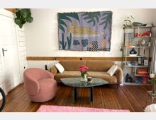 Wohnzimmer mit Holzboden, einer beigen Couch und einem rosafarbenen Sessel. Zentral steht ein grüner Marmortisch mit einer Vase voller rosa Blumen, darüber hängt ein Wandteppich mit abstraktem Pflanzen- und Figurenmotiv. Im Hintergrund ein weißes Bücherregal mit farbigen Büchern und Dekoration sowie eine Zimmerpflanze.