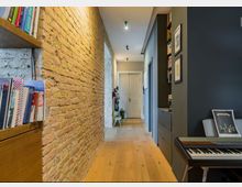 Ein schmaler, modern eingerichteter Flur mit einer freiliegenden Backsteinwand auf der linken Seite, Holzboden und weißen Zimmertüren im Hintergrund. Auf der rechten Seite befinden sich dunkle Wände mit Schränken, Bildern und einem E-Piano.