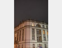 Ein beleuchtetes historisches Gebäude bei Nacht mit klassizistischen Architekturmerkmalen, darunter dekorative Reliefs, mehrere symmetrisch angeordnete Fenster und ein kleiner Balkon mit Balustrade. Teile der Fassade sind angestrahlt, und der wolkenverhangene Himmel bildet den Hintergrund.