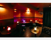 Innenraum einer Lounge- oder Bar-Sitzecke mit gepolsterter roter Rundbank, kleinen runden Tischen und gedimmten Tischlampen. Orangefarbene Wände mit Wandleuchten, dunkler Fliesenboden und ein schwarzer Vorhang an der rechten Seite prägen den Raum.