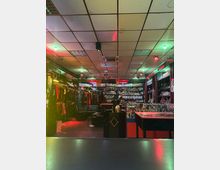 Ein Verkaufsraum mit niedriger Rasterdecke, bunter Beleuchtung und Schaufensterpuppen. Regale an den Wänden präsentieren Kleidung und Accessoires, während Glasvitrinen kleine Artikel ausstellen.