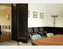 Wartezimmer mit vier gepolsterten Stühlen an einer weißen Wand, drei gerahmten Bildern darüber und einem schwarzen Metall-Kleiderständer; ein Holztisch ist im Vordergrund, und eine offene Tür führt zu einem angrenzenden Raum mit Holzvertäfelung.