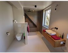 Modernes Badezimmer mit bodengleicher Walk-in-Dusche mit kleinen dunkelgrauen Fliesen und wandhängender Toilette hinter einer halbhohen Trennwand. Rechts ein schwebender Holz-Waschtisch mit ovalem Aufsatzbecken und schwarzer Armatur neben einem großen Fenster.