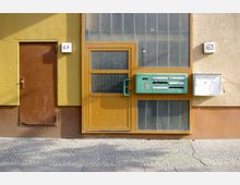 Gelbe Hausfassade mit einer braunen Tür und einer danebenliegenden Eingangstür aus Glas und Holzrahmen. An der Wand befinden sich grüne Briefkästen und zwei Hausnummernschilder mit der Nummer 63.