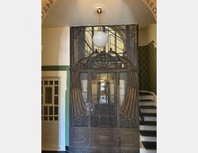 Eingangsbereich in einem historischen Gebäude mit einer verzierten Metalltür, die dekorative Ornamente und Reliefs zeigt. Im Hintergrund sind eine Wendeltreppe mit Holzgeländer und ein Kronleuchter mit kugelförmiger Lampe sichtbar.