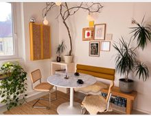 Helle Ecke eines Wohnraums mit einem runden Marmortisch, Stühlen mit Geflecht, einer gelben Polsterbank und Pflanzen. An der Wand hängen gerahmte Bilder, darüber eine minimalistische Hängelampe mit Ästen, neben einem großen Fenster mit Blick nach draußen.