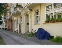 Straßenansicht eines Wohngebäudes im Altbaustil mit hellgelber Fassade, großen Fenstern und kunstvoll verziertem Eingangsportal. Vor dem Haus stehen Fahrräder, eines davon an die Wand gelehnt, und auf den Fensterbänken befinden sich Blumenkästen mit grünen und blühenden Pflanzen.