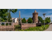 Historische Szene mit einem runden, aus Backstein erbauten Turm mit Zinnen, einer Brücke aus Stein mit Geländer und einem Denkmal mit einem Obelisken und einer Statue. Im Vordergrund fließt ein Fluss, umgeben von Vegetation, im Hintergrund befinden sich Bäume und Wohnhäuser.