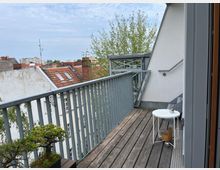 Balkon mit Holzboden und Metallgeländer, ausgestattet mit einem kleinen weißen Beistelltisch und einer dekorativen Pflanze. Im Hintergrund sind Dächer von Wohngebäuden sowie ein Baum und ein leicht bewölkter Himmel zu sehen.