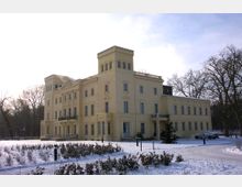 Gelbes historisches Gebäude im Schlossstil mit rechteckigen Türmen und symmetrischen Fenstern, umgeben von schneebedecktem Garten und Bäumen im Winter. Ein verschneiter Weg führt zum Gebäude.