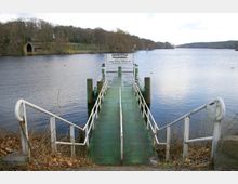 Metallsteg mit Geländern führt zu einem Anleger auf einem ruhigen See, mit der Beschilderung „Anlegestelle Pfaueninsel“. Im Hintergrund ist ein bewaldetes Ufer mit einem kleinen Bootshaus zu sehen.