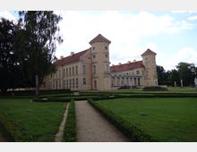 Schlossgebäude mit beigefarbener Fassade und roten Ziegeldächern, flankiert von zwei Rundtürmen. Im Vordergrund ein formaler Garten mit symmetrischen Wegen, Hecken und Rasenflächen, umgeben von Bäumen und Skulpturen.