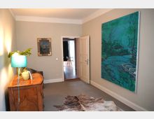 Ein schmaler Wohnraum mit beigem Teppichboden, einer Holzkommode mit einer grünen Tischlampe und einer Zimmerpflanze. An den Wänden hängen zwei Bilder: ein gerahmtes Gemälde und ein großes, abstraktes Landschaftsgemälde in Blau- und Grüntönen; durch eine geöffnete Tür ist ein weiteres Zimmer mit Holzboden und einem Stuhl sichtbar.