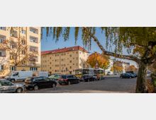 Straßenszene in einem Wohngebiet mit mehrstöckigen gelb-beigen Gebäuden und roten Dächern, parkenden Autos und einem Bus. Im Vordergrund ist ein Baum mit herabhängenden Ästen und herbstlich gefärbte Bäume zu sehen.