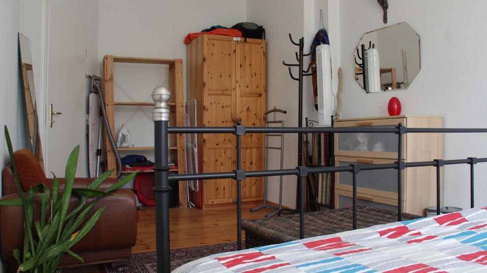 Interior of a bedroom with a black metal bedframe in the foreground, wooden wardrobe and open shelving against white walls, and a dresser with an octagonal wall mirror. The room has a hardwood floor with a patterned rug, a brown chair, a potted plant, and several coat racks and stored household items.