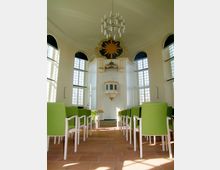 Innenraum einer kleinen Kapelle mit hellen Wänden und großen Rundbogenfenstern. Im Vordergrund stehen grüne Stühle, während im hinteren Bereich ein erhöhter Holzkanzelaltar mit einer goldenen Strahlensonne an der Wand sichtbar ist.