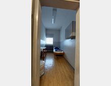 Kleiner Raum mit schlichten Möbeln, darunter ein Einzelbett, ein kleiner Tisch und ein Waschbecken. Der Raum hat helle Wände, einen Holzboden und ein vergittertes Fenster; eine Toilette ist links im Vordergrund sichtbar.