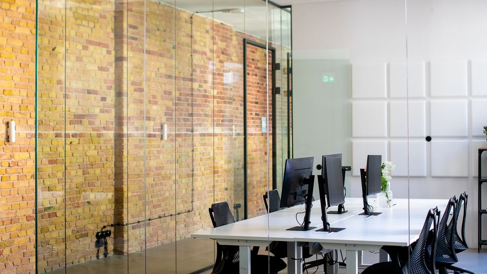 Glasverkleideter Besprechungsraum in einem modernen Büro mit freiliegender Ziegelwand, langem weißen Tisch und mehreren Schreibtischstühlen mit Monitoren. An der Decke sind rechteckige Lichtpaneele und ein beleuchtetes Notausgangsschild sichtbar.