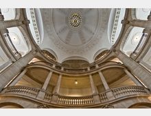 Innenansicht einer historischen Kuppelhalle mit verzierten Rundbögen, säulengetragenen Galerieebenen und einer reich dekorierten Kuppeldecke mit zentralem Kronleuchter. Die Architektur besteht aus hellem Stein und weist klassische Stilelemente auf.