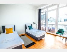 Helles Schlafzimmer mit zwei Einzelbetten, weißer Bettwäsche und farbigen Kissen, Holzboden und großem Fenster, das auf einen Balkon mit Sitzmöbeln und Blick auf moderne Gebäude und Wasser führt. Ein grüner Stuhl und ein kleiner Tisch stehen rechts im Raum.