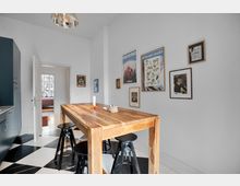 Moderne Küche mit einem rechteckigen Holztisch und schwarzen Hockern auf einem schwarz-weiß karierten Boden. An der weißen Wand hängen verschiedene gerahmte Bilder, links befindet sich eine blaue Küchenzeile, und eine Tür führt in einen angrenzenden Raum mit großen Fenstern.