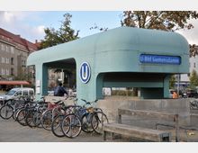 U-Bahneingang der Station "Siemensdamm" in Berlin mit einer türkisfarbenen Überdachung, auf der das U-Bahn-Symbol und der Stationsname zu sehen sind. Im Vordergrund sind zahlreiche geparkte Fahrräder sowie eine Holzbank, während Wohnhäuser und Bäume die Umgebung prägen.