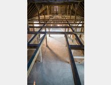 Innenansicht eines Dachbodens mit offenliegender Holzkonstruktion. Sichtbar sind Schrägdachbalken, gemauerte Wände aus Ziegeln und ein Betonboden, durchbrochen von Licht, das durch kleine Fensteröffnungen einfällt.