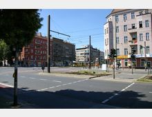 Straßenecke in einer Stadt mit mehrstöckigen Wohn- und Geschäftsgebäuden, darunter ein rotes und ein rosafarbenes Gebäude. Im Vordergrund sind Straßenbahngleise, Verkehrsschilder und Ampeln sichtbar, während im Hintergrund eine Straßenbahn und Fahrradfahrer die Szene ergänzen.