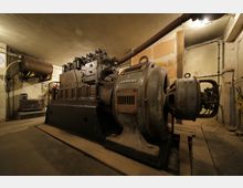 Ein alter Maschinenraum mit einer massiven Siemens-Maschine, bestehend aus einem Generator und mechanischen Komponenten. Der Raum ist schlicht gestaltet, mit Betonwänden, freiliegenden Rohren und gedämpfter Beleuchtung.