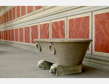 Ein großer Steintrog mit rechteckiger Form, seitlichen Metallgriffen und Löwenköpfen als Sockeldekoration steht in einem Flur mit rot marmorierten Wandpaneelen und klassischen weißen Rahmen. Der Boden ist mit Steinfliesen ausgelegt.