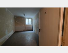 Ein leerer Raum mit unbehandelten Wänden, Betonestrichboden und einem kleinen Fenster mit Heizkörper darunter. Eine Tür mit der Nummer „3“ steht offen und gewährt den Blick in den Raum.