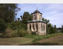 Kleiner historischer Pavillon aus Stein mit Rundbogenfenstern und verzierten Säulen, umgeben von dichtem Grün und einer Wiese; im Hintergrund sind Bäume und ein Gewässer zu sehen.