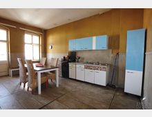 Ein einfacher Küchenraum mit gelb gestrichenen Wänden, einer Küchenzeile mit weißen Unterschränken und blauen Oberschränken sowie einem freistehenden Kühlschrank. Im Vordergrund steht ein Esstisch aus Holz mit vier Flechtstühlen, die auf einem gefliesten Boden angeordnet sind.