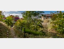 Ein schmaler, mit Blättern bedeckter Weg führt durch eine grüne, von Bäumen und Sträuchern bewachsene Landschaft. Im Hintergrund ist ein roter Regionalzug auf einer erhöhten Bahnstrecke sowie ein Gebäude mit Ziegeldach zu sehen.