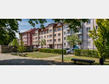 Eine kleine Parkanlage mit gepflastertem Weg, Rasenflächen, Bänken und Bäumen im Vordergrund. Im Hintergrund stehen mehrstöckige Wohngebäude mit Balkonen und roten Dächern, sowie parkende Autos entlang der Straße.