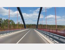 Straßenbrücke mit zwei großen, blauen Stahlbögen und roten Seilen, die die Fahrbahn überspannen. Im Hintergrund ist ein dichter Nadelwald unter einem leicht bewölkten Himmel zu sehen.