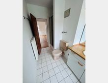 Schmales Badezimmer mit weiß gefliesten Bodenfliesen, WC und Waschtisch mit Holzplatte sowie Spiegelschrank. Links befindet sich ein Heizkörper und eine geöffnete Tür führt in einen angrenzenden Raum mit Holzfußboden.