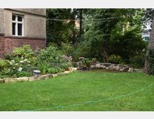 Ein grüner Garten mit einer Rasenfläche, blühenden Pflanzen und Sträuchern sowie einer Steinumrandung. Im Hintergrund steht ein Gebäude mit einer Fassade aus Backstein und Putz, umgeben von großen Bäumen und dichter Vegetation.