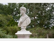 Eine klassische Marmorbüste eines Mannes mit gelocktem Haar und einem Umhang, auf einem Sockel stehend, vor einer Kulisse aus dichtem, grünem Laub und hohen Bäumen.