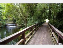 Holzbrücke mit Geländer über einen kleinen Fluss in einer waldartigen Umgebung, umgeben von dichtem grünen Laub. Im Hintergrund ist ein kleiner Wasserfall und ein weiteres Brückengeländer sichtbar.