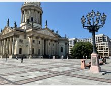 Historisches Gebäude mit Säulen und Figuren auf einem großen, gepflasterten Platz, vermutlich der Deutsche Dom in Berlin. Im Vordergrund eine verzierte Laterne, im Hintergrund moderne Gebäude und ein Baum.