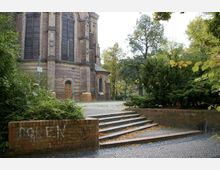 Teilansicht einer historischen Kirche aus Backstein mit großen, spitzbogenförmigen Fenstern und verzierten Fassadenelementen. Im Vordergrund befindet sich eine Treppe mit niedrigen Backsteinmauern, umgeben von dichtem Grün und Bäumen.