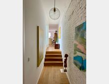 Flur mit Holzboden und weißen Wänden, auf der rechten Seite eine weiß gestrichene Ziegelwand mit farbigen Gemälden und einer kleinen Holzskulptur. Im Hintergrund führt eine kleine Holztreppe zu einem Raum mit Möbeln und Fensterlicht.