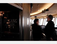 Ein Innenraum mit großen Fenstern, die Tageslicht hereinlassen, dekoriert mit Blumenarrangements an der Decke. Im Vordergrund ist eine Tür mit der Aufschrift „Ausgang“ und „WC“ zu sehen, während zwei Personen miteinander sprechen.