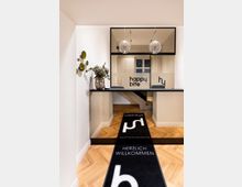 Modernes Empfangsbereich mit hellem Holzfußboden, einem schwarzen Tresen und einer Glasscheibe mit dem Logo „happy bite“. Zwei kugelförmige Deckenleuchten hängen über dem Tresen, links steht eine Blumenvase, und ein Teppich mit der Aufschrift „Herzlich Willkommen“ liegt in der Mitte des Raumes.