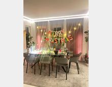 Essbereich in einem modernen Raum mit einem gedeckten Tisch, umgeben von acht grauen Polsterstühlen, dekoriert mit Weihnachtslichtern und einem „Merry Christmas“-Bannerschriftzug vor bodentiefen Jalousien. Pflanzen und dezente Beleuchtung ergänzen das Ambiente.