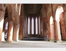 Innenraum einer historischen gotischen Kirche mit hohen Spitzbogenfenstern, roten Backsteinmauern und verzierten Säulen. Ein hölzernes Dachgerüst ist sichtbar, und im Hintergrund befindet sich eine kleine erhöhte Bühne.