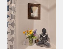 Stein-Buddha-Statue mit einer weißen Perlenkette, daneben ein Glas mit bunten Blumen (Gänseblümchen, Ringelblumen und Lavendel) auf einer hellen Fliesen-Oberfläche. Im Hintergrund hängt ein antik wirkender Holzrahmenspiegel, links ist ein Vorhang mit blau-weißem geometrischem Muster sichtbar.